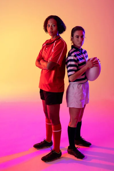 Portrait of two diverse female rugby players with rugby ball over neon pink lighting. Sport, movement, training and active lifestyle concept.