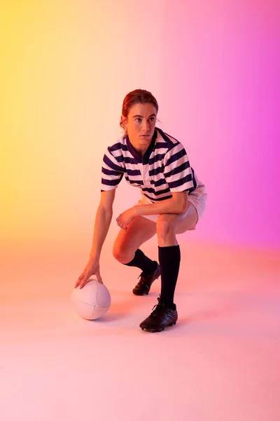 Caucasian female rugby player crouching with rugby ball over neon pink lighting. Sport, movement, training and active lifestyle concept.