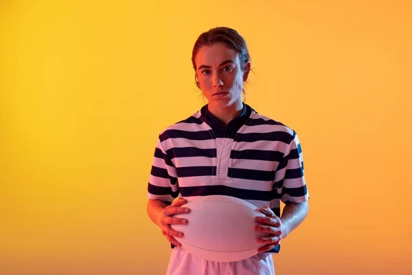 Portrait of caucasian female rugby player with rugby ball over neon yellow lighting. Sport, movement, training and active lifestyle concept.