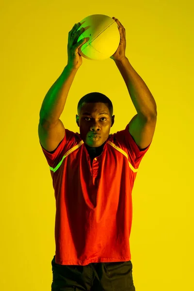 Portrait of african american male rugby player with rugby ball over yellow lighting. Sport, movement, training and active lifestyle concept.