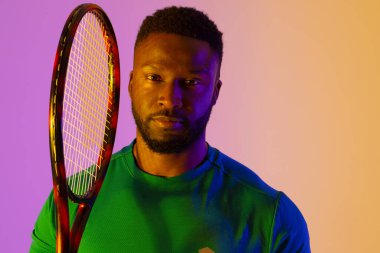 Image of african american male tennis player in violet and yellow neon lighting. Sport, movement, fitness and active lifestyle concept.