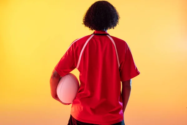 Rear view of african american female rugby player with rugby ball over neon yellow lighting. Sport, movement, training and active lifestyle concept.