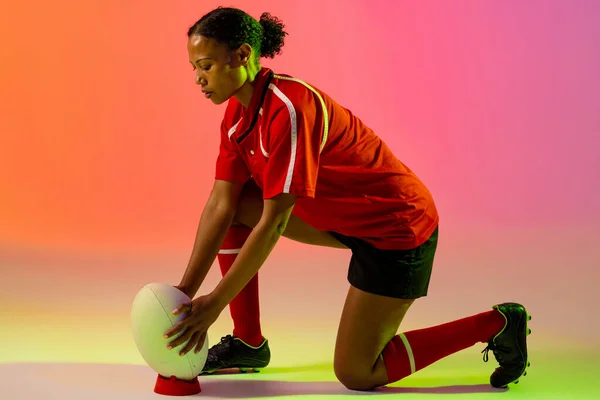 African american female rugby player crouching with rugby ball over neon pink lighting. Sport, movement, training and active lifestyle concept.