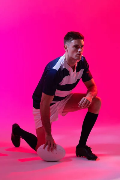 Caucasian male rugby player crouching with rugby ball over pink lighting. Sport, movement, training and active lifestyle concept.