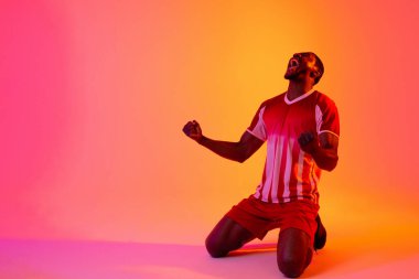 Happy african american male soccer player raising hands over neon pink lighting. Sport, movement, training and active lifestyle concept.
