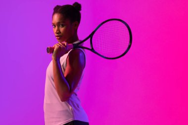 Image of african american female tennis player in violet and pink neon lighting. Sport, movement, fitness and active lifestyle concept.