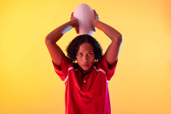 Portrait of african american female rugby player with rugby ball over neon yellow lighting. Sport, movement, training and active lifestyle concept.