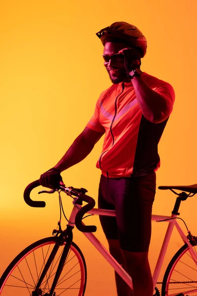 Pionowy Obraz Szczęśliwego Afrykańskiego Rowerzysty Patrzącego Kamerę Neonowym Pomarańczowym Oświetleniu — Zdjęcie stockowe