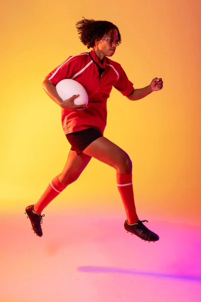 African american female rugby player running with rugby ball over neon pink lighting. Sport, movement, training and active lifestyle concept.