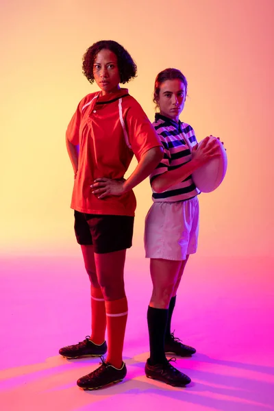 Portrait of two diverse female rugby players with rugby ball over neon pink lighting. Sport, movement, training and active lifestyle concept.
