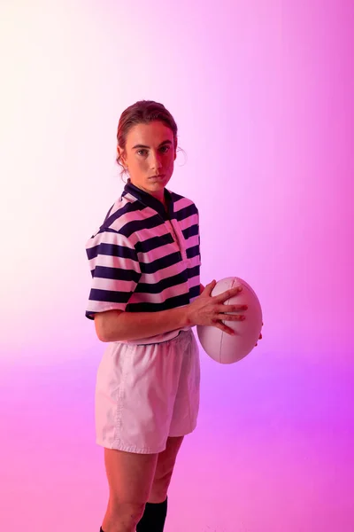 Portrait of caucasian female rugby player with rugby ball over neon pink lighting. Sport, movement, training and active lifestyle concept.