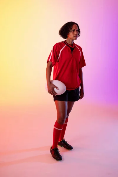 African american female rugby player with rugby ball over neon pink lighting. Sport, movement, training and active lifestyle concept.