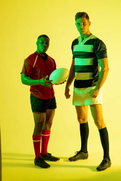 Portrait of diverse male rugby players with rugby ball over yellow lighting. Sport, movement, training and active lifestyle concept.