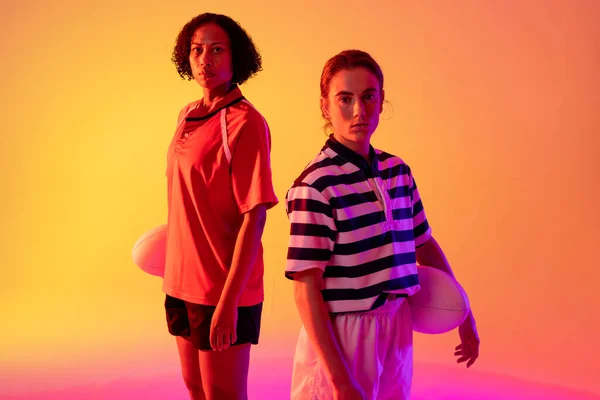 Portrait of diverse female rugby players with rugby balls over neon pink lighting. Sport, movement, training and active lifestyle concept.