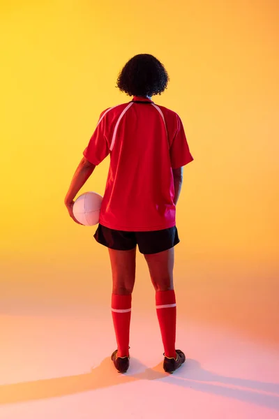 Rear view of african american female rugby player with rugby ball over neon yellow lighting. Sport, movement, training and active lifestyle concept.