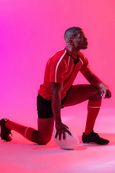 African american male rugby player crouching with rugby ball over pink lighting. Sport, movement, training and active lifestyle concept.