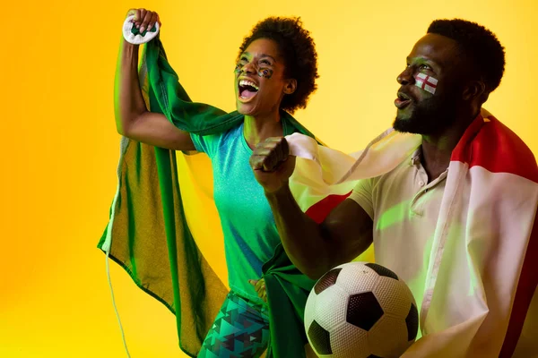 Image of african american soccer fan couple with flags of brazil and england in yellow lighting. Sport, fans, cheering and emotions concept.