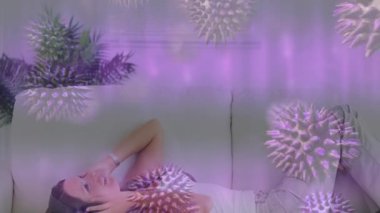 Kafkasyalı bir kadının evdeki kanepede yatarken kulaklık taktığı covid-19 hücrelerinin birleşik videosu. Covid-19 salgın konsepti