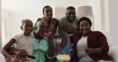 Animation of ball and team colours over happy african american family watching match. Sport, cheering and spending time together concept digitally generated video.