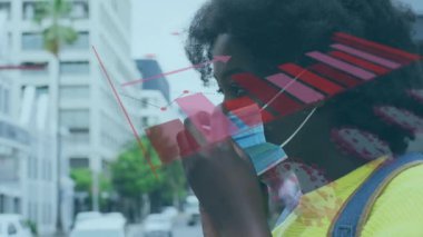 Animation of virus cells and graphs over african american woman in face mask. Communication and technology during covid 19 pandemic concept digitally generated video.