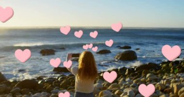 Animation of hearts over back view of caucasian woman taking photo on beach. Holidays, vacations, relax, social media and global communication concept digitally generated video.