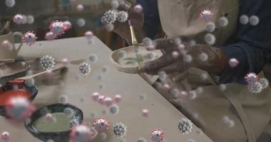 Animation of falling covid 19 cells over african american potter. Global covid 19 pandemic, science and connections concept digitally