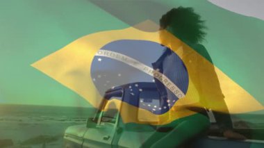 Animation of brazilian flag waving over african american woman sitting on pick-up truck at beach. Digital composite, multiple exposure, nature, vacation, patriotism, identity concept.