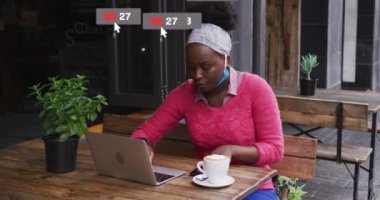 Animation of social media icons over african american woman wearing face mask. Global covid 19 pandemic, social media, data processing and digital interface concept digitally generated video.