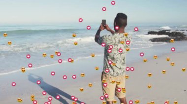 Animation of heart and love emojis over african american man taking smartphone photos on sunny beach. Global communication network, holidays and social media concept digitally generated video.