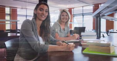 Animation of globe and programming language over smiling caucasian teachers sitting in school. Digital composite, multiple exposure, globalization, database, office, education, technology concept.