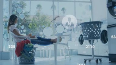 Bilgisayar simgeleriyle çamaşırhanede beklerken laptopta çalışan melez bir kadının videosu. Dijital kompozit, kolaylık, çamaşırhane, bağlantı, küresel iletişim, veri, ağ.