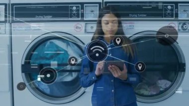 Çamaşırhanede ve iletişim ikonlarında melez genç bir kadının tablet bilgisayar kullanmasının birleşik videosu. Teknoloji, kolaylık, çamaşırhane, grafik, yaşam tarzı, bağlantı, ağ oluşturma, küresel iletişim.