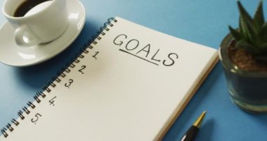 Video of notebook with goals and copy space, coffee and pencil on blue surface. Business, working in office, equipment and tools concept.