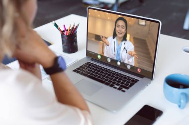 Sırıtkan Asyalı kadın doktorla dizüstü bilgisayar görüşmesi yapan bir kadının omuz manzarası. Tıbbi hizmetler, teletıp ve dijital arayüz kavramı.