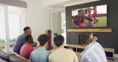 Kanepede oturan ve evdeki televizyonda futbol izleyen farklı arkadaşların videosu. Spor, rekabet ve ailece evde vakit geçirmek..