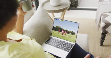 Çift ırklı bir kadının kanepede oturup dizüstü bilgisayarda futbol izlediği bir video. Spor, rekabet ve ailece evde vakit geçirmek..