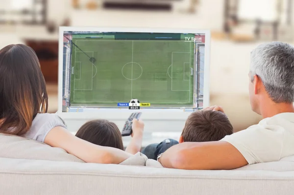 Family sitting on sofa watching television together against football players tacakling for the ball on pitch