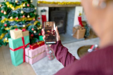 Kadın, Noel Baba 'nın maskeli haliyle akıllı telefon videosu çekiyor. Covid 19 salgını sırasında Noel, şenlik ve iletişim teknolojisi.