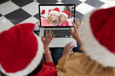 Noel Baba şapkalı beyaz anne ve çocuk arkadaşlarıyla birlikte dizüstü bilgisayardan yılbaşı görüntülerini izliyorlar. Noel, şenlik ve iletişim teknolojisi.