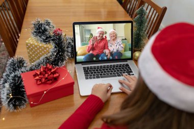 Kafkasyalı bir kadın mutlu bir şekilde kıdemli ailelerine el sallayarak dizüstü bilgisayarla Noel videosu çekiyor. Noel, şenlik ve iletişim teknolojisi.