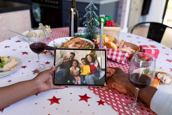 Hands of african american couple holding tablet with african american family on screen. christmas, festivity and communication technology during coronavirus covid 19 pandemic.