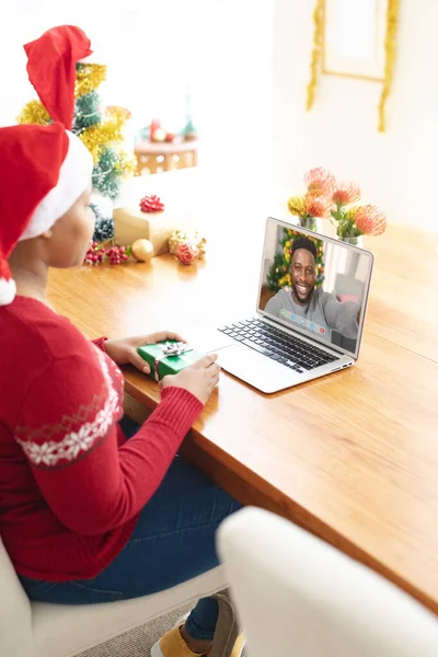 サンタの帽子のアフリカ系アメリカ人女性は 笑顔の男性の友人とラップトップのクリスマスビデオ通話を作ります クリスマスやお祭りや通信技術は — ストック写真
