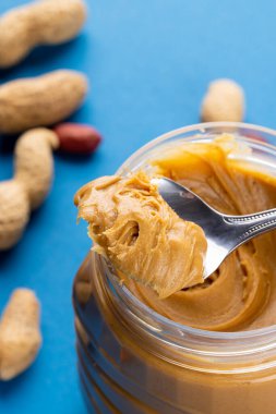 Mavi yüzeyde fıstık ezmeli kaşık ve kavanozun dikey görüntüsü. Yiyecek, kahvaltı, beslenme ve Amerikan mutfağı konsepti.