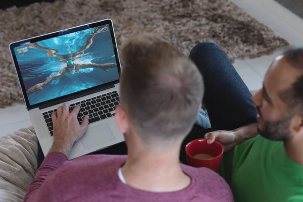 Two diverse male friends at home watching swimming competition on laptop. sports, competition, entertainment and technology concept digital composite image.