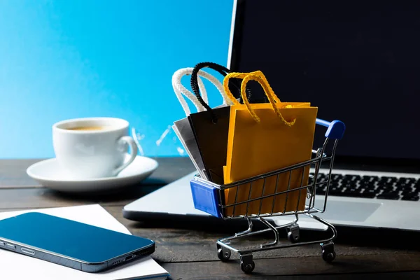 Composition of shopping cart with bags and laptop on blue background. Retail, shopping and black friday concept.