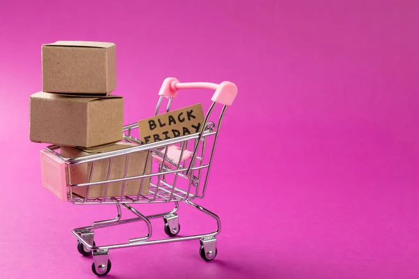 Composition of shopping cart with boxes and black friday text on pink background. Retail, shopping and black friday concept.