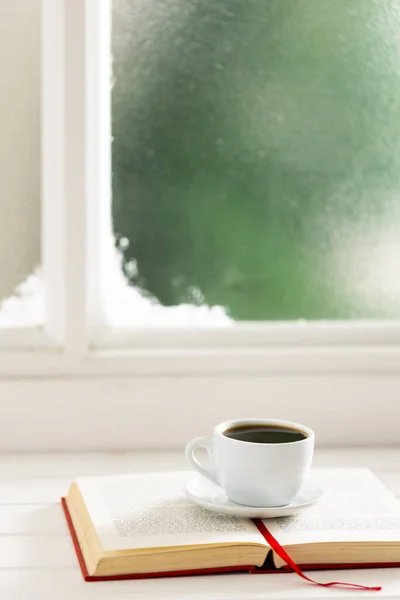 Cup of hot coffee and book over frozen and snowy window. Christmas, tradition and celebration concept.