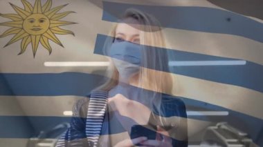 Uruguay bayrağının şehir caddesinde yüz maskesi takan beyaz bir kadının üzerinde dalgalanması. covid 19 salgın konsepti boyunca dijital olarak üretilen küresel sağlık önlemleri.