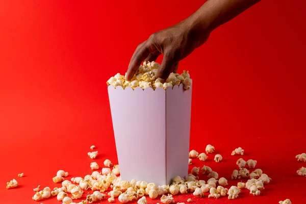 Image of hand of african american man taking away pop corn from box on red surface. Food, snacks, cinema and american cuisine concept.