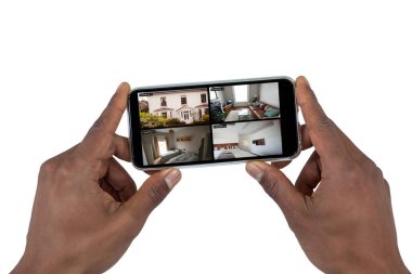 Beyaz perdedeki güvenlik kameralarından evi gören akıllı telefonu tutan Afro-Amerikan bir adamın elleri. güvenlik, güvenlik, izleme ve dijital arayüz.